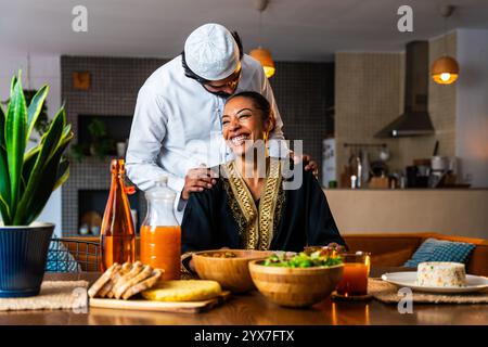 Glückliches, schönes, junges arabisches Paar mit emiratischer Kleidung, das zu Hause zusammen zu Mittag essen - Mann und Frau aus dem Nahen Osten in traditioneller arabischer Kleidung Stockfoto