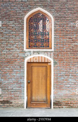 Außenwand der Juma-Moschee, Ganja, Aserbaidschan: Rustikale rote Backsteinwand mit wunderschön gefertigter Holztür an der Basis und einem aufwendig gestalteten bogenförmigen Fenster darüber, das kulturelle Kunstfertigkeit zeigt Stockfoto