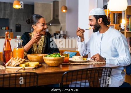 Glückliches, schönes, junges arabisches Paar mit emiratischer Kleidung, das zu Hause zusammen zu Mittag essen - Mann und Frau aus dem Nahen Osten in traditioneller arabischer Kleidung Stockfoto