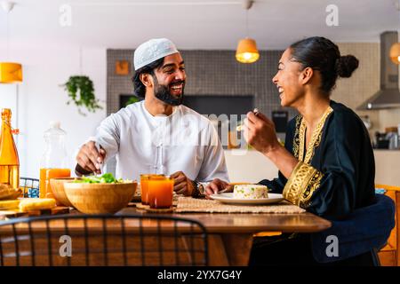 Glückliches, schönes, junges arabisches Paar mit emiratischer Kleidung, das zu Hause zusammen zu Mittag essen - Mann und Frau aus dem Nahen Osten in traditioneller arabischer Kleidung Stockfoto
