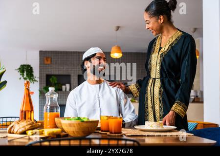 Glückliches, schönes, junges arabisches Paar mit emiratischer Kleidung, das zu Hause zusammen zu Mittag essen - Mann und Frau aus dem Nahen Osten in traditioneller arabischer Kleidung Stockfoto