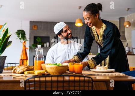 Glückliches, schönes, junges arabisches Paar mit emiratischer Kleidung, das zu Hause zusammen zu Mittag essen - Mann und Frau aus dem Nahen Osten in traditioneller arabischer Kleidung Stockfoto