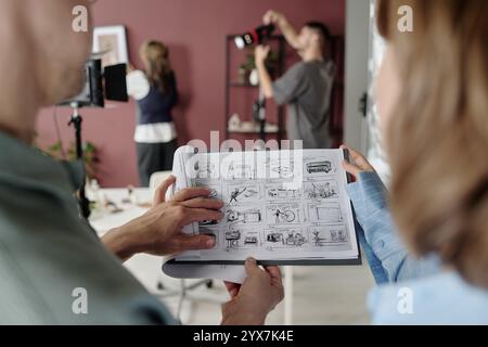 Hochwinkelaufnahme von nicht erkennbaren Männern und Frauen, die Schwarzweißskizzen auf Papier betrachten, während die Crew in unscharfem Hintergrund arbeitet Stockfoto