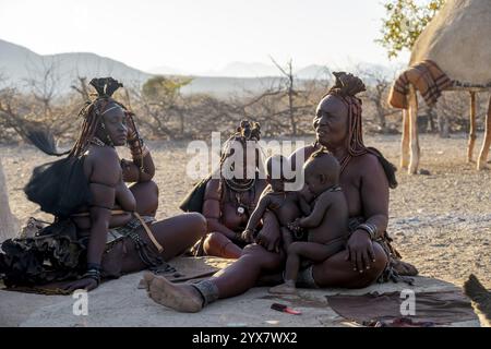 Sie heiratete Himba-Frau mit ihren Babys in den Armen, die vor der Hütte der ersten Frau saßen, morgens im traditionellen Himba-Dorf Kaokove Stockfoto