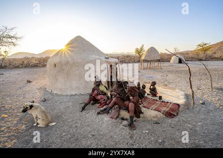 Himba Frau und Kinder sitzen vor der ersten Frauenhütte, Sonnenaufgang, Sonnenstern, traditionelles Himba Dorf, Kaokoveld, Kunene, Namibia, Afrika Stockfoto