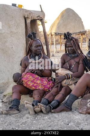 Sie heiratete Himba-Frau mit ihren Babys in den Armen, die vor der Hütte der ersten Frau saßen, morgens im traditionellen Himba-Dorf Kaokove Stockfoto