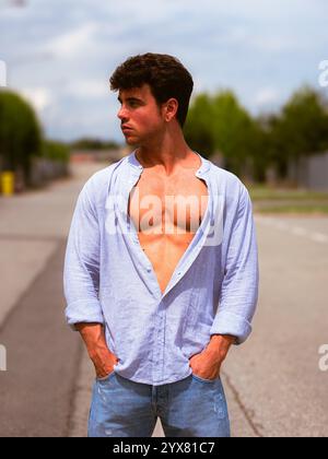 Ein junger Mann steht selbstbewusst auf einer leeren Straße, trägt ein hellblaues offenes Hemd und Jeans. Die Sonne erleuchtet seinen straffen Körper, während die Bäume säumen Stockfoto