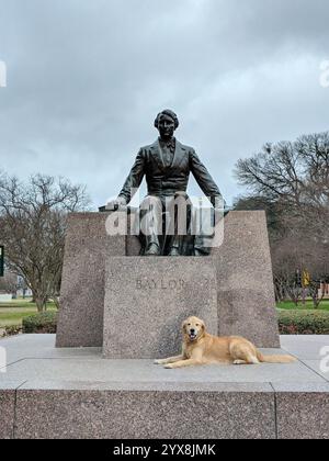 Ein Golden Retriever sitzt ruhig am Fuße einer Statue von Richter Baylor auf dem Campus der Baylor University in Waco, Texas. Stockfoto