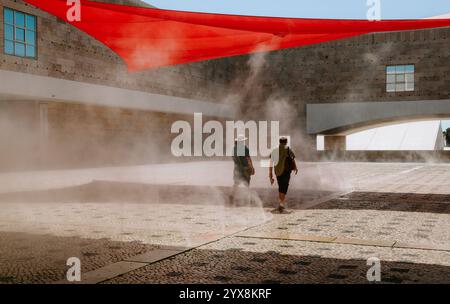 Zwei Personen genießen erfrischenden Nebel und spazieren durch den Innenhof des Centro Cultural de belem in lissabon unter leuchtendem roten Schatten Stockfoto