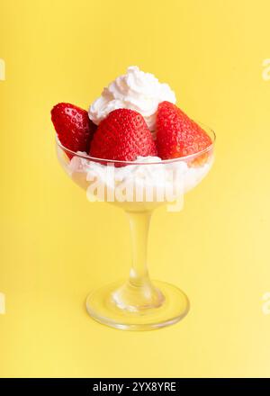 Frische Erdbeeren und Schlagsahne, serviert in einem Glas auf hellgelbem Hintergrund, die Süße, Frische und eine sommerliche Dessertatmosphäre verkörpern. Stockfoto