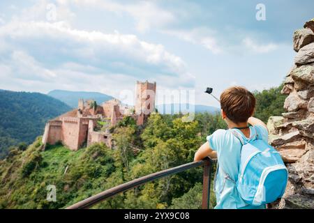 Ein kleiner Junge mit blauem Rucksack macht ein Selfie, während er die Ruinen eines alten Schlosses bewundert, umgeben von üppigen grünen Hügeln in Europa, Elsass, Stockfoto