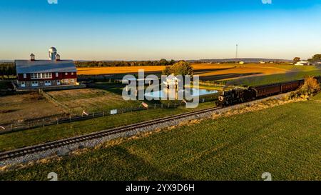 Die historische Dampfeisenbahn fährt entlang der Gleise, vorbei an einer malerischen roten Scheune, ausgedehnten Ackerland. Die Sonne geht unter und wirft warme Goldtöne über die Landschaft. Stockfoto