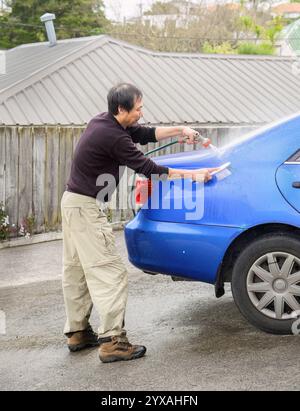 Mann, der das Auto auf der Auffahrt mit einer Bürste und einem Wasserspray aus einem Schlauch wäscht. Vertikales Format. Stockfoto