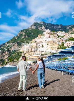Ein vielseitiges Paar genießt einen romantischen Spaziergang am Kiesstrand von Positano, Italien, umgeben von lebhaften Häusern und üppigen Bergen. Die Sonne scheint hell und verschönert die atemberaubende Küste. Stockfoto