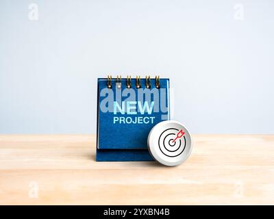 Modernes Ziel-Pfeil-Bullseye-Symbol auf blauem Schreibtischkalenderdeckel mit Text „NEW PROJECT“ auf Holztisch, weißer Hintergrund. Geschäftsziel, Erfolgsziel sc Stockfoto