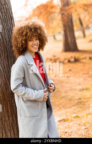 Eine junge Frau mit lockigen Haaren lehnt sich an einen Baum, trägt einen warmen Pullover, lächelt fröhlich im goldenen Herbstlaub und umhergefallenen Blättern Stockfoto