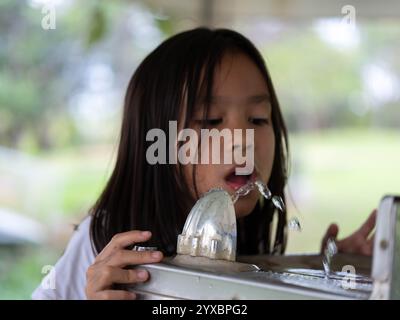 Das süße Mädchen trinkt Wasser aus einem quadratischen, glänzenden Metallbrunnen in einem Park. Kinder trinken Wasser aus einem öffentlichen Wasserspender im Freien. Stockfoto