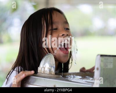 Das süße Mädchen trinkt Wasser aus einem quadratischen, glänzenden Metallbrunnen in einem Park. Kinder trinken Wasser aus einem öffentlichen Wasserspender im Freien. Stockfoto