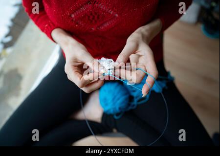 Nahaufnahme der Hände, die kunstvoll mit blauem Garn stricken und sich auf komplizierte Nähte konzentrieren, vor einem gemütlichen Hintergrund mit einem roten Pullover und einem Garnknäuel Stockfoto