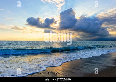 Ein Segelboot bei Sonnenuntergang sitzt auf dem Ozean mit Sail Open, während Sonnenstrahlen durch die Wolken brechen Stockfoto