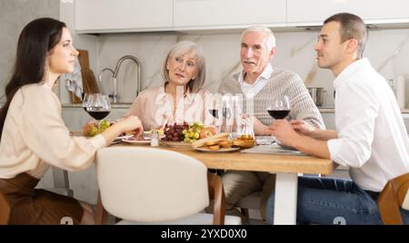 Fröhliche Seniorinnen und Senioren genießen ein Abendessen mit einem jungen Paar zu Hause Stockfoto