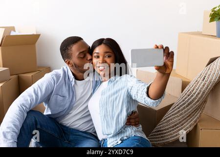 Glückliche schwarze Ehepartner machen Selfie in ihrer neuen Wohnung, feiern den Umzugstag, sitzen in der Nähe von Kartonkisten Stockfoto