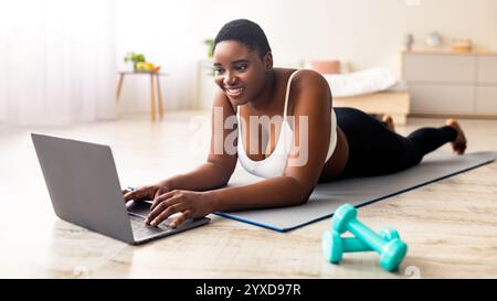 Kurvige schwarze Frau, die Online-Sportvideos auf einem Laptop ansieht, auf dem Boden liegt und vor dem Heimtraining ein Gadget benutzt Stockfoto