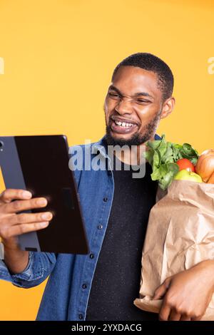 Fröhlicher, fröhlicher Erwachsener schaut auf seinem modernen Tablet in die sozialen Medien und bringt eine Tasche mit Bio-Lebensmitteln aus dem lokalen Supermarkt. afroamerikaner, fröhlicher Mensch, schaut sich Gadget im Studio an. Stockfoto