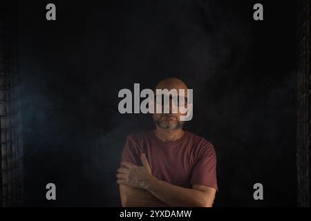 Ein kahler und bärtiger Mann, der eine Brille trägt und in die Kamera schaut. Isoliert auf einem rauchigen schwarzen Hintergrund. Südamerika Stockfoto