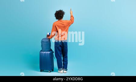 Junger Mann signalisiert, während er mit Koffer auf blauem Hintergrund steht Stockfoto
