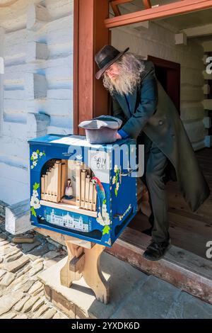Sanok, Polen - 4. Mai 2024. Ein bärtiger Straßenmusiker mit Hut und Mantel steht an der Tür eines Holzhauses und spielt eine handbemalte Tonnorgel. Stockfoto