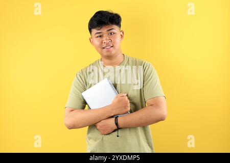 Junger asiatischer Mann, der Tablet-Computer hält, mit grünem Avocado T-Shirt, isoliert auf gelbem Hintergrund Stockfoto