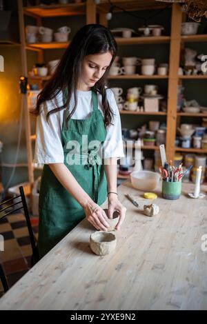Herstellung von handwerklicher Keramik. Arbeitstisch im Keramikstudio mit Frau, die nassen Tonbecher formt Stockfoto