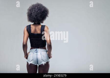 Rückansicht einer jungen schwarzen Frau mit Afro-Frisur, die selbstbewusst mit Händen auf der Hüfte posiert. Mit Jeansshorts und stylischem grauen Rücken Stockfoto