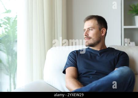Selbstbewusster Mann, der durch ein Fenster schaut und zu Hause auf einem Sofa sitzt Stockfoto