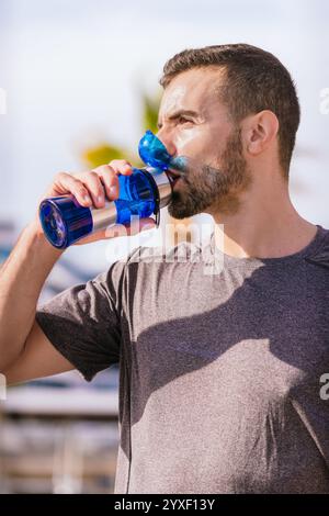 Sportlicher Mann trinkt Wasser aus der roten Flasche draußen im Stadtpark. Vertikal Stockfoto