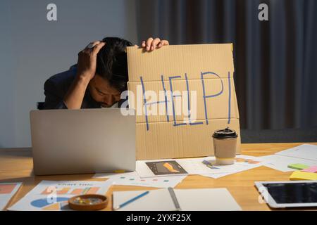 Frustrierter und überarbeiteter Geschäftsmann, der seinen Kopf mit einem Laptop hält und nachts im Büro um Hilfe bittet. Stockfoto