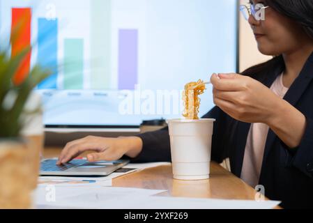 Glückliche asiatische Geschäftsfrau muss Nudeln essen, während sie im Büro am Laptop arbeitet. Seitenansicht, selektiver Fokus. Stockfoto