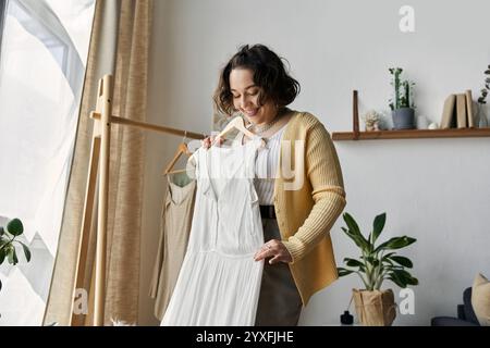 Eine junge Frau mit lockigen Haaren schaut sich in ihrem lebendigen Wohnraum gerne ein Kleid an. Stockfoto