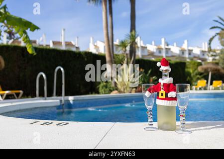 Foto einer Sektflasche mit einem kleinen Weihnachtsmann-Outfit, umgeben von zwei Weingläsern an einem sonnigen Tag am Swimmingpool, der Weihnachten anzeigt Stockfoto