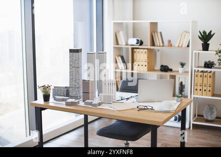 Moderner Bürotisch mit Architekturmodell und Laptop-Einrichtung. Der helle Arbeitsbereich umfasst Pflanzen, Akten und Brillen. Ideal für die Konzeption von Stadtplanungsprojekten mit zeitgenössischen Elementen. Stockfoto