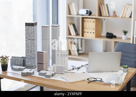Moderner Bürotisch mit Architekturmodell und Laptop-Einrichtung. Der helle Arbeitsbereich umfasst Pflanzen, Akten und Brillen. Ideal für die Konzeption von Stadtplanungsprojekten mit zeitgenössischen Elementen. Stockfoto