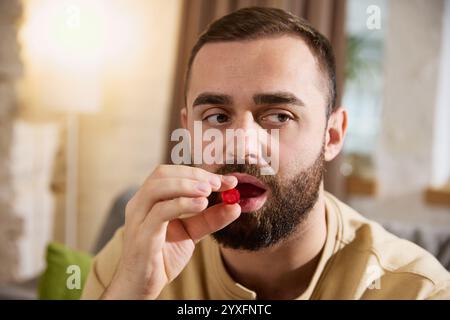 Nahaufnahme eines jungen, kranken Mannes in lässigem Outfit, der an Halsschmerzen leidet, trinkt rote Lutschtabletten, sitzt in einem hellen, gemütlichen Wohnzimmer. Stockfoto