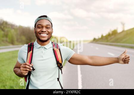 Lächelnder schwarzer Kerl mit Rucksack, der das Auto heruntergetrampelt, die freie Fahrt mitnimmt, auf der Autobahn anhängt, freier Platz Stockfoto