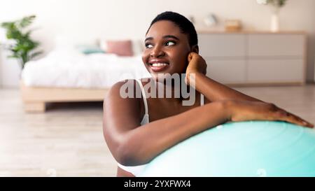 Fröhliche übergewichtige schwarze Frau, die sich auf den Fitnessball stützt und in Innenräumen Sport macht Stockfoto