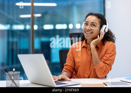 Zielgerichtete junge Frau lächelt und hört Musik über Kopfhörer mit Laptop. Sie sitzt an einem modernen Büroarbeitsplatz und scheint in ihre Arbeit eingetaucht zu sein. Stockfoto