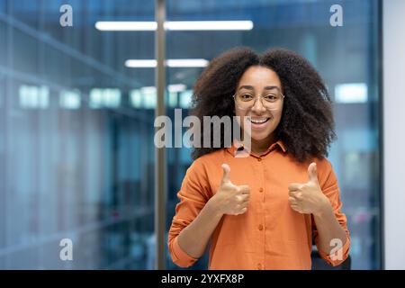 Fröhliche junge Frau mit Brille und lockigem Haar trägt ein orangenes Hemd. Sie lächelt herzlich und zeigt sich in einer modernen Büroumgebung doppelt hoch und bringt Positivität und Motivation zum Ausdruck Stockfoto