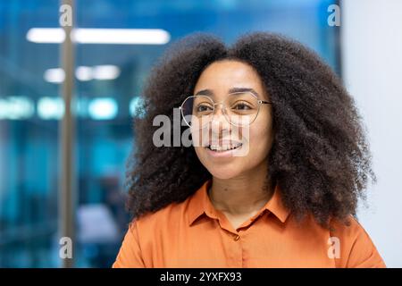 Eine selbstbewusste Frau mit lockigen Haaren und Brillen, lässig gekleidet, lächelt in einem modernen Büro. Stockfoto