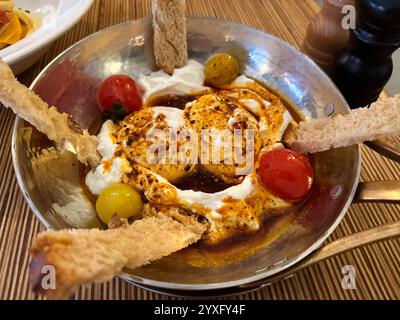 Türkische Eier oder Cilbir werden mit griechischem Joghurt kombiniert mit frischen Kräutern und Knoblauch in der Nähe auf dem Teller auf dem Holztisch zubereitet Stockfoto