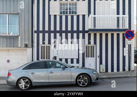 Costa Nova, Portugal - 11. September 2024 : Ein zweistöckiges gestreiftes Haus steht neben modernen Gebäuden mit geparkten Autos in der Nähe. Stockfoto